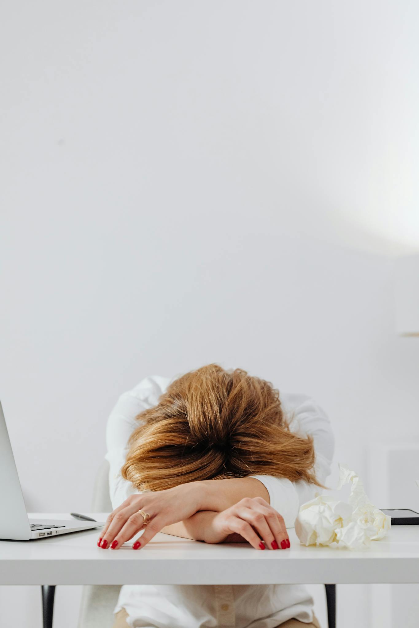 A tired woman with red nails resting her head on a white desk in a bright office setting.